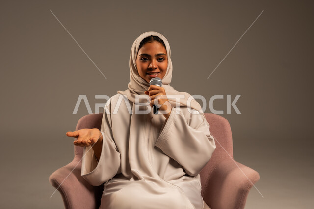 مفهوم الفن وتأثيره الايجابي على الامرأة، تقديم العروض الفنيه و الغنائيه والفقرات الترفيهية، بدء التدريب الصوتي، بورتريه لشابة عربية خليجية سعودية محجبة ترتدي العباءة الملونة وتجلس على كرسي مريح تمسك بيدها المايكروفون بإيماءات الاندماج بالغناء، خلفيه رمادية