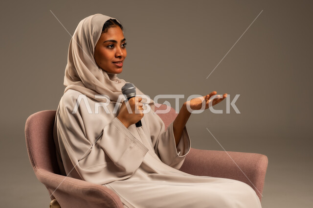 مفهوم الفن وتأثيره الايجابي على الامرأة، تقديم العروض الفنيه و الغنائيه والفقرات الترفيهية، بدء التدريب الصوتي، بورتريه لشابة عربية خليجية سعودية محجبة ترتدي العباءة الملونة وتجلس على كرسي مريح تمسك بيدها المايكروفون بإيماءات الاندماج بالغناء، خلفيه رمادية
