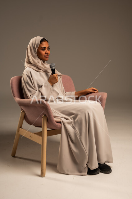 مفهوم الفن وتأثيره الايجابي على الامرأة، تقديم العروض الفنيه و الغنائيه والفقرات الترفيهية، بدء التدريب الصوتي، بورتريه لشابة عربية خليجية سعودية محجبة ترتدي العباءة الملونة وتجلس على كرسي مريح تمسك بيدها المايكروفون بإيماءات الاندماج بالغناء، خلفيه رمادية