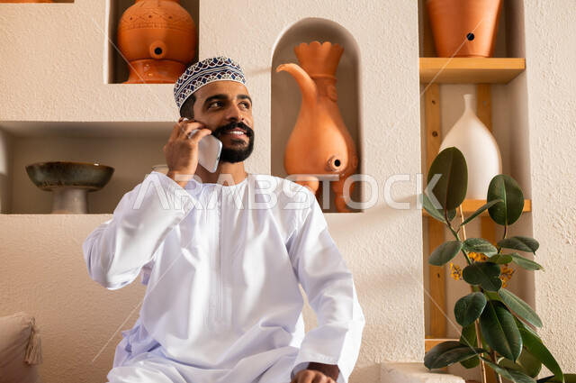 الرد على الاتصالات والمكالمات, صورة مقربة لرجل عربي خليجي عماني يرتدي الدشداشة والكمه يجلس في محل لبيع الهدايا التذكارية التقليدية يقوم بإجراء مكالمة هاتفية عبر الجوال، التواصل مع الأهل والأصدقاء عبر الهاتف المحمول، استخدام الأجهزة الحديثة والمتطورة، 