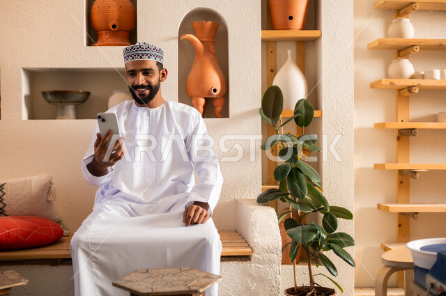 استخدام الأجهزة الحديثة والمتطورة، الرد على الاتصالات والمكالمات, صورة مقربة لرجل عربي خليجي عماني يرتدي الدشداشة والكمه يجلس في محل لبيع الهدايا والتحف يقوم بالنظر إلى الموبايل، التواصل مع الأهل والأصدقاء عبر الهاتف المحمول