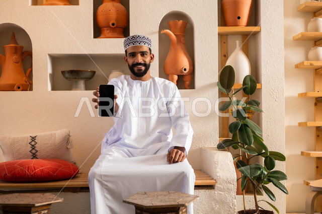 Communicating with family and friends via mobile phone, using modern and advanced devices, answering communications and calls, a close-up photo of an Arab, Gulf, Omani man wearing a dishdasha and a sleeve, sitting in a shop selling gifts and antiques, displaying a black screen on his mobile phone.