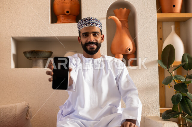 التواصل مع الأهل والأصدقاء عبر الهاتف المحمول، استخدام الأجهزة الحديثة والمتطورة، الرد على الاتصالات والمكالمات, صورة مقربة لرجل عربي خليجي عماني يرتدي الدشداشة والكمه يجلس في محل لبيع الهدايا والتحف يقوم بعرض شاشة سوداء عبر الجوال