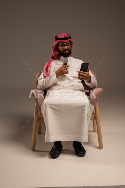 القراءة من الهاتف المحمول، القيام بالحوارات والنقاشات، بورتريه لرجل عربي خليجي سعودي يرتدي الثوب التقليدي والشماغ يجلس على الكرسي يحمل بيده مكبر الصوت ويقوم بإلقاء الخطابات، استخدام الأجهزة التقنية الحديثة، خلفية رمادية