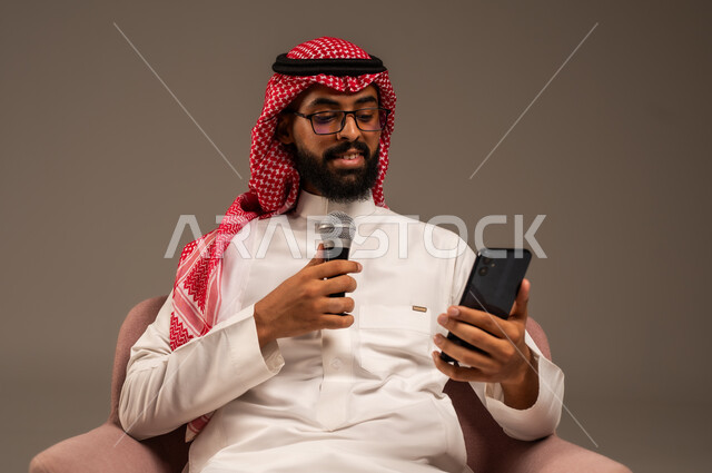 استخدام الأجهزة التقنية الحديثة، القراءة من الهاتف المحمول، القيام بالحوارات والنقاشات، بورتريه مقرب لرجل عربي خليجي سعودي يرتدي الثوب التقليدي والشماغ يجلس على الكرسي يحمل بيده مكبر الصوت ويقوم بإلقاء الخطابات، خلفية رمادية