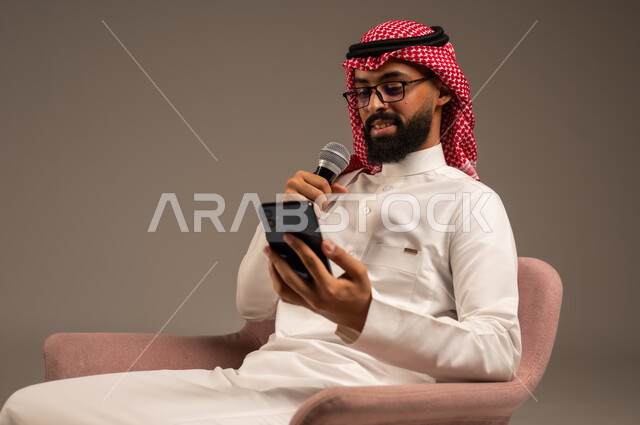 القراءة من الهاتف المحمول، القيام بالحوارات والنقاشات، بورتريه مقرب لرجل عربي خليجي سعودي يرتدي الثوب التقليدي والشماغ يجلس على الكرسي يحمل بيده مكبر الصوت ويقوم بإلقاء الخطابات، استخدام الأجهزة التقنية الحديثة، خلفية رمادية