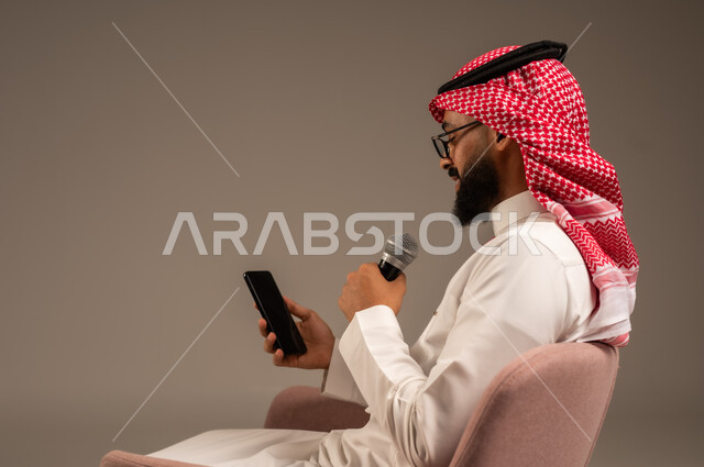 القيام بالحوارات والنقاشات، بورتريه مقرب لرجل عربي خليجي سعودي يرتدي الثوب التقليدي والشماغ يجلس على الكرسي يحمل بيده مكبر الصوت ويقوم بإلقاء الخطابات، القراءة من الهاتف المحمول، استخدام الأجهزة التقنية الحديثة، خلفية رمادية