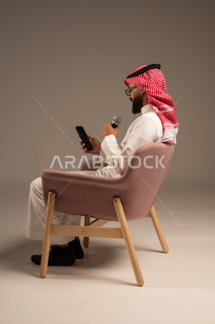 Conducting dialogues and discussions, portrait of a Saudi Arabian Gulf man wearing traditional thobe and shemagh sitting on a chair holding a loudspeaker and giving speeches, reading from a mobile phone, using modern technical devices, gray background