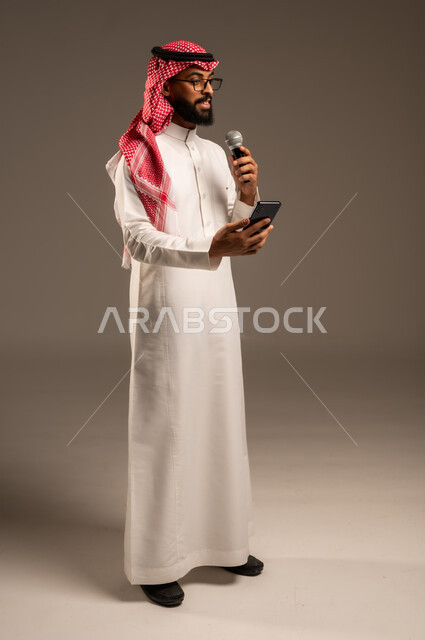 Conducting dialogues and discussions, portrait of a Saudi Arabian Gulf man wearing traditional thobe and shemagh holding a mobile phone and loudspeaker giving speeches and looking at something, using modern technical devices, full body image, gray background