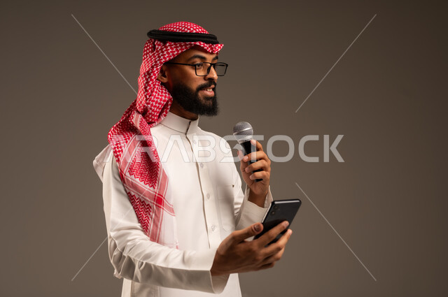 القيام بالحوارات والنقاشات، بورتريه مقرب لرجل عربي خليجي سعودي يرتدي الثوب التقليدي والشماغ يحمل بيده هاتف محمول ومكبر الصوت يقوم بإلقاء الخطابات وينظر إلى شيء ما، استخدام الأجهزة التقنية الحديثة، خلفية رمادية
