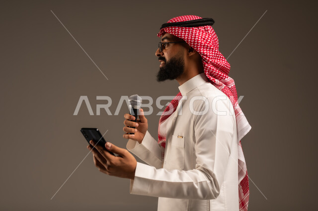 القيام بالحوارات والنقاشات، بورتريه مقرب من الجانب لرجل عربي خليجي سعودي يرتدي الثوب التقليدي والشماغ يحمل بيده هاتف محمول ومكبر الصوت يقوم بإلقاء الخطابات وينظر إلى شيء ما، استخدام الأجهزة التقنية الحديثة، خلفية رمادية