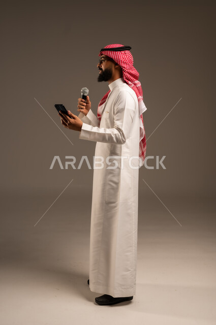 القيام بالحوارات والنقاشات، بورتريه من الجانب لرجل عربي خليجي سعودي يرتدي الثوب التقليدي والشماغ يحمل بيده هاتف محمول ومكبر الصوت يقوم بإلقاء الخطابات وينظر إلى شيء ما، استخدام الأجهزة التقنية الحديثة، صورة الجسم بالطول الكامل، خلفية رمادية