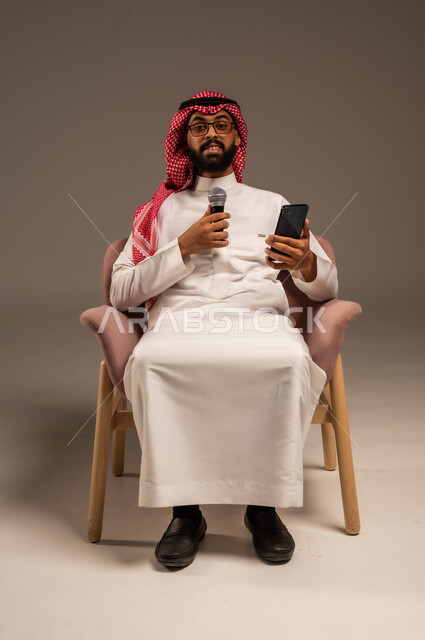 استخدام الأجهزة التقنية الحديثة، القيام بالحوارات والنقاشات، بورتريه لرجل عربي خليجي سعودي يرتدي الثوب التقليدي والشماغ يجلس على الكرسي يحمل بيده هاتف محمول ومكبر الصوت يقوم بإلقاء الخطابات وينظر إلى الكاميرا، خلفية رمادية