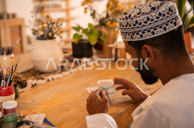 احتراف تلوين الخزف والفخاريات، محترف عربي خليجي عماني يرتدي الدشداشة والكمه يعمل في ورشة الاعمال الفنية، ممارسة مهارة صنع الفخار، أعمال يدوية عمانية، صوره مقربة لحرفي يقوم بالنقش على الأواني الفخار