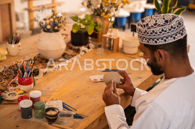 احتراف تلوين الخزف والفخاريات، محترف عربي خليجي عماني يرتدي الدشداشة والكمه يعمل في ورشة الاعمال الفنية، ممارسة مهارة صنع الفخار، أعمال يدوية عمانية، صوره مقربة لحرفي يقوم بالنقش على الأواني الفخار