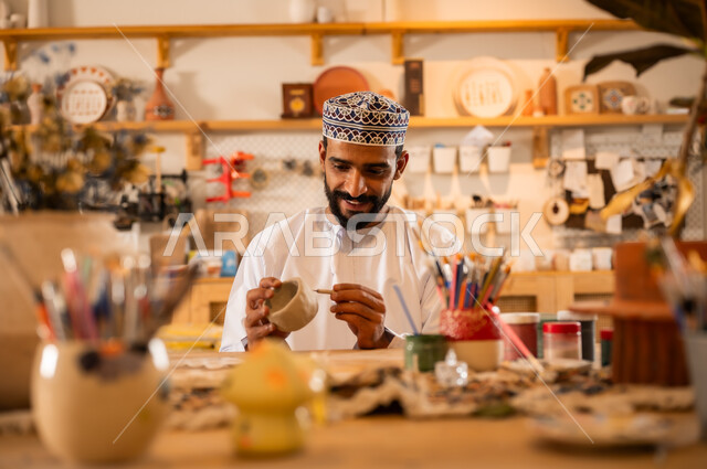 احتراف تلوين الخزف والفخاريات، محترف عربي خليجي عماني يرتدي الدشداشة والكمه يعمل في ورشة الاعمال الفنية، ممارسة مهارة صنع الفخار، أعمال يدوية عمانية، صوره مقربة لحرفي يقوم بالنقش على الأواني الفخار