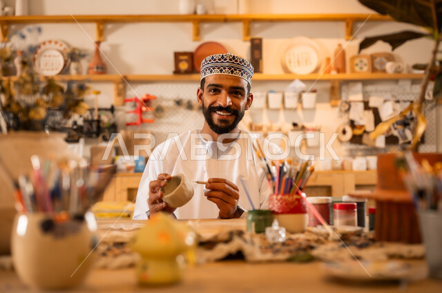 احتراف تلوين الخزف والفخاريات، محترف عربي خليجي عماني يرتدي الدشداشة والكمه يعمل في ورشة الاعمال الفنية، ممارسة مهارة صنع الفخار، أعمال يدوية عمانية، صوره مقربة لحرفي يقوم بالنقش على الأواني الفخار