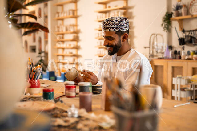 احتراف تلوين الخزف والفخاريات، محترف عربي خليجي عماني يرتدي الدشداشة والكمه يعمل في ورشة الاعمال الفنية، ممارسة مهارة صنع الفخار، أعمال يدوية عمانية، صوره مقربة لحرفي يقوم بالنقش على الأواني الفخار