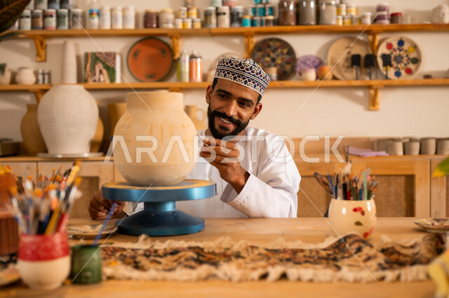 احتراف تلوين الخزف والفخاريات، محترف عربي خليجي عماني يرتدي الدشداشة والكمه يعمل في ورشة الاعمال الفنية، ممارسة مهارة صنع الفخار، أعمال يدوية عمانية، صوره مقربة لحرفي يقوم بالنقش على الأواني الفخار