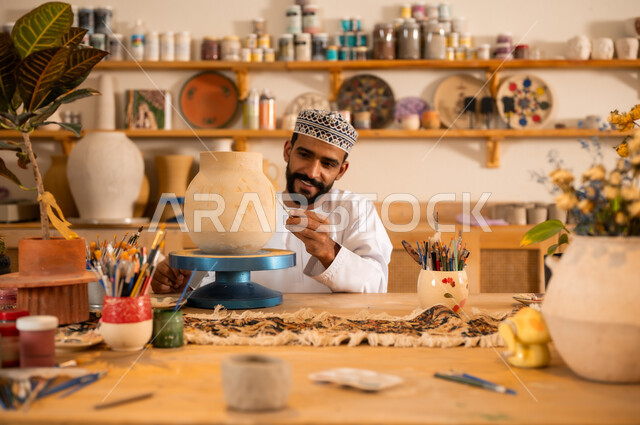 احتراف تلوين الخزف والفخاريات، محترف عربي خليجي عماني يرتدي الدشداشة والكمه يعمل في ورشة الاعمال الفنية، ممارسة مهارة صنع الفخار، أعمال يدوية عمانية، صوره مقربة لحرفي يقوم بالنقش على الأواني الفخار