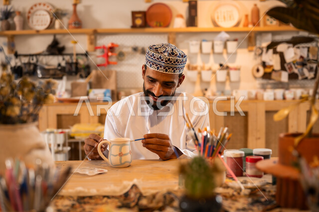 أعمال يدوية عمانية، احتراف تلوين الخزف والفخاريات، محترف عربي خليجي عماني يرتدي الدشداشة والكمه يعمل في ورشة الاعمال الفنية، ممارسة مهارة صنع الفخار،  صوره مقربة لحرفي يقوم بالنقش على الفخار