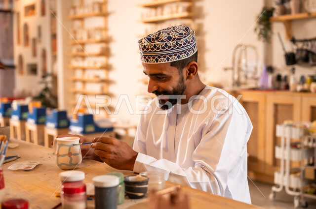 أعمال يدوية عمانية، احتراف تلوين الخزف والفخاريات، محترف عربي خليجي عماني يرتدي الدشداشة والكمه يعمل في ورشة الاعمال الفنية، ممارسة مهارة صنع الفخار،  صوره مقربة لحرفي يقوم بالنقش على الفخار