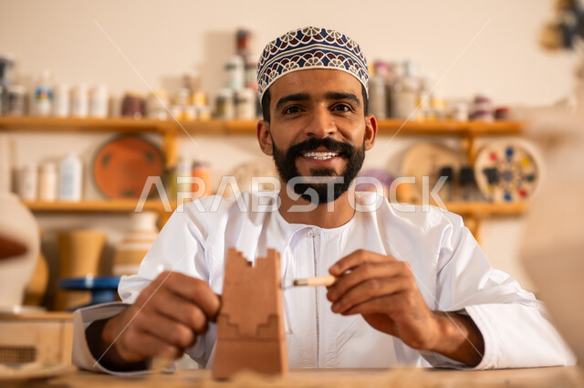أعمال يدوية عمانية، احتراف تلوين الخزف والفخاريات، محترف عربي خليجي عماني يرتدي الدشداشة والكمه يعمل في ورشة الاعمال الفنية، ممارسة مهارة صنع الفخار،  صوره مقربة لحرفي يقوم بالنقش على الفخار