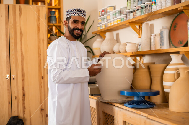 محترف عربي خليجي عماني يرتدي الدشداشة والكمه يعمل في ورشة الاعمال الفنية، احتراف تلوين الخزف والفخاريات، ممارسة مهارة صنع الفخار، أعمال يدوية عمانية، صوره مقربة لحرفي يقوم بالنقش على الأواني الفخار