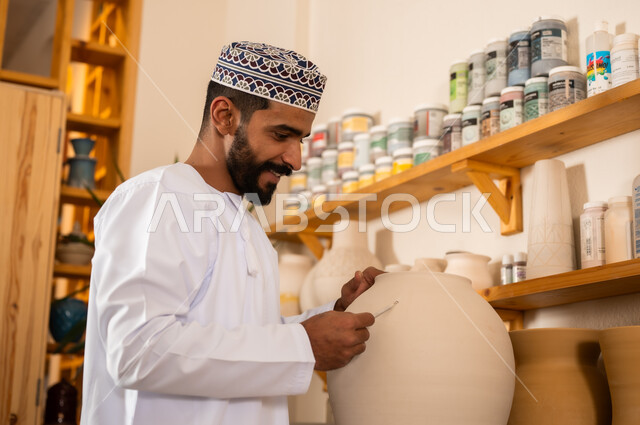 محترف عربي خليجي عماني يرتدي الدشداشة والكمه يعمل في ورشة الاعمال الفنية، احتراف تلوين الخزف والفخاريات، ممارسة مهارة صنع الفخار، أعمال يدوية عمانية، صوره مقربة لحرفي يقوم بالنقش على الأواني الفخار