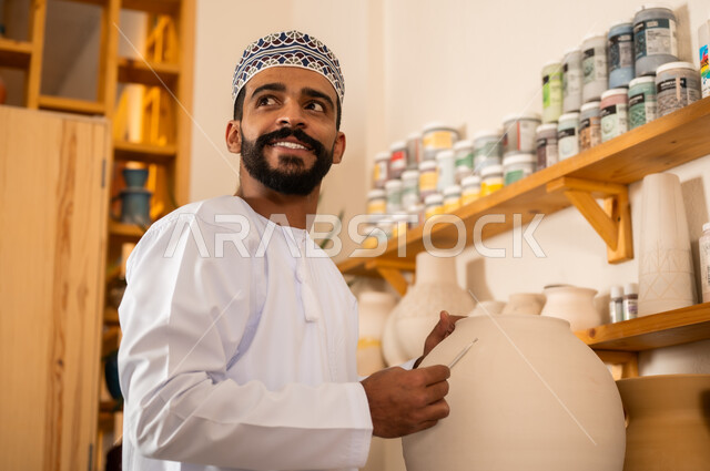 Professional Arab Gulf Omani man wearing dishdasha and kummah working in art workshop, professional pottery and ceramics coloring, practicing pottery making skill, Omani handicrafts, close up of craftsman engraving on pottery