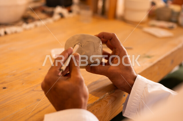 احتراف تلوين الخزف والفخاريات، محترف عربي خليجي عماني يرتدي الدشداشة والكمه يعمل في ورشة الاعمال الفنية، ممارسة مهارة صنع الفخار، أعمال يدوية عمانية، صوره مقربة لحرفي يقوم بالنقش على الأواني الفخار