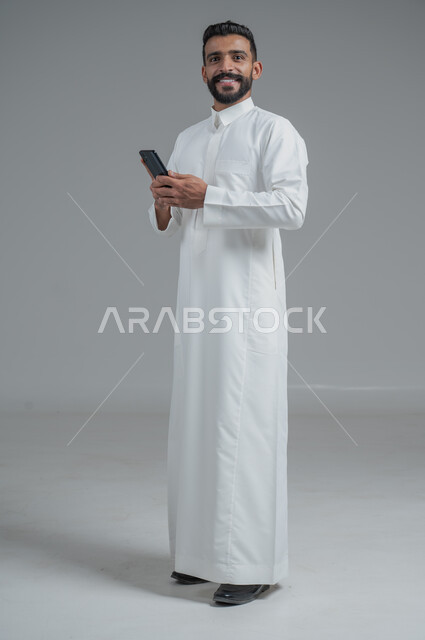 Communicating with family and friends via mobile phone, using modern and advanced devices, answering calls and calls, portrait of a Saudi Arabian Gulf man wearing traditional dress holding a mobile phone in his hand, full-length body image, gray background