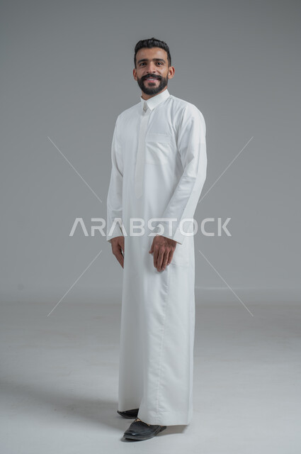 الوقوف بثقة واستقامة، الاهتمام بالمظهر الخارجي والأناقة، مفهوم الرجولة والثقة بالنفس، بورتريه لرجل عربي خليجي سعودي مبتسم يرتدي الثوب التقليدي ينظر الى الكاميرا، صورة الجسم بالطول الكامل، خلفية رمادية
