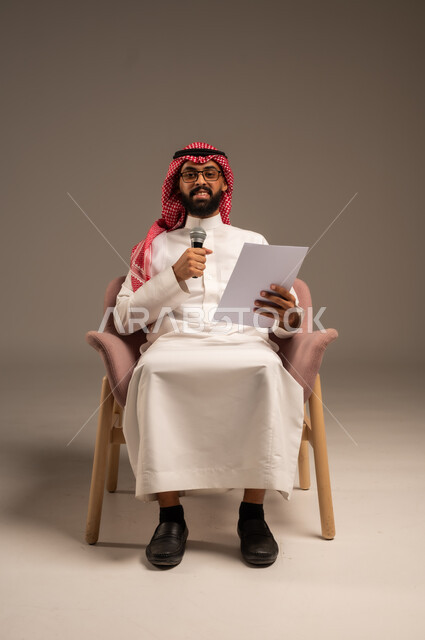 قراءة الملاحظات والمعلومات، بورتريه لرجل عربي خليجي سعودي يرتدي الثوب التقليدي والشماغ يجلس على الكرسي يحمل بيده مجموعة من الأوراق ومكبر الصوت يقوم بإلقاء الخطابات وينظر إلى الكاميرا، إعداد وتحضير أفكار الخطاب بشكل مسبق، خلفية رمادية