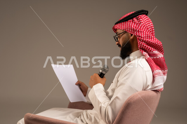 قراءة الملاحظات والمعلومات، القيام بالحوارات والنقاشات، بورتريه مقرب من الجانب لرجل عربي خليجي سعودي يرتدي الثوب التقليدي والشماغ يجلس على الكرسي يحمل بيده مجموعة من الأوراق ومكبر الصوت ويقوم بإلقاء الخطابات، إعداد وتحضير أفكار الخطاب بشكل مسبق، خلفية رمادية
