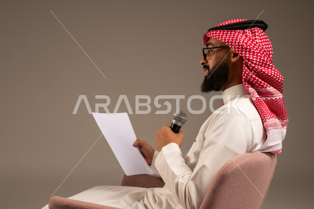 القيام بالحوارات والنقاشات، بورتريه مقرب من الجانب لرجل عربي خليجي سعودي يرتدي الثوب التقليدي والشماغ يجلس على الكرسي يحمل بيده مجموعة من الأوراق ومكبر الصوت يقوم بإلقاء الخطابات، قراءة الملاحظات والمعلومات، إعداد وتحضير أفكار الخطاب بشكل مسبق، خلفية رمادية