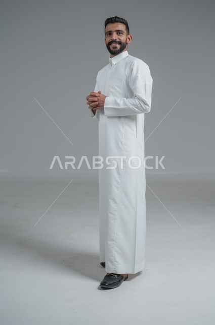 الوقوف بثقة واستقامة، الاهتمام بالمظهر الخارجي والأناقة، مفهوم الرجولة والثقة بالنفس، بورتريه لرجل عربي خليجي سعودي مبتسم يرتدي الثوب التقليدي ينظر الى الكاميرا يقف بايدي متشابكة، صورة الجسم بالطول الكامل، خلفية رمادية