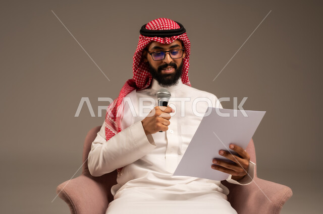 قراءة الملاحظات والمعلومات، بورتريه مقرب لرجل عربي خليجي سعودي يرتدي الثوب التقليدي والشماغ يجلس على الكرسي يحمل بيده مجموعة من الأوراق ومكبر الصوت يقوم بإلقاء الخطابات، القيام بالحوارات والنقاشات، إعداد وتحضير أفكار الخطاب بشكل مسبق، خلفية رمادية