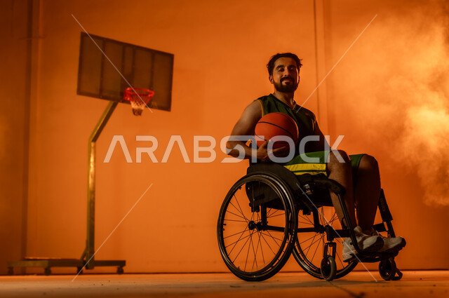 التدرب على إتقان لعب كرة السلة، أنشطة ترفيهية ورياضات شبابية، تحسين حياة ذوي الإعاقة الحركية، المحافظة على اللياقة البدنية والليونة، بورتريه لشاب عربي خليجي سعودي يرتدي الملابس الرياضية يجلس على كرسي متحرك يمسك الكرة بيده، اتباع نمط حياة صحي، خلفية برتقالية