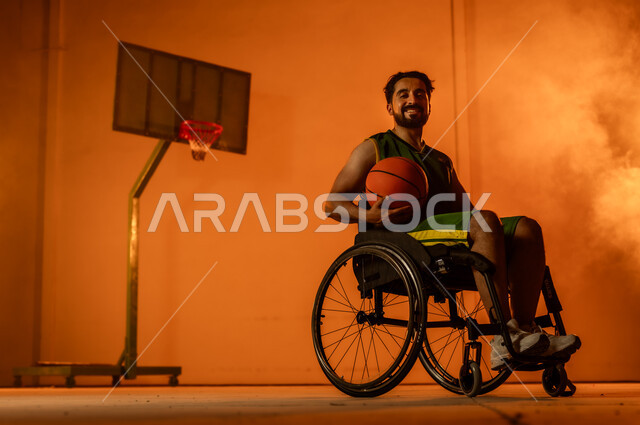 التدرب على إتقان لعب كرة السلة، أنشطة ترفيهية ورياضات شبابية، تحسين حياة ذوي الإعاقة الحركية، المحافظة على اللياقة البدنية والليونة، بورتريه لشاب عربي خليجي سعودي يرتدي الملابس الرياضية يجلس على كرسي متحرك يمسك الكرة بيده، اتباع نمط حياة صحي، خلفية برتقالية
