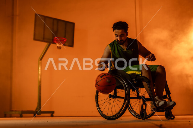 التدرب على إتقان لعب كرة السلة، أنشطة ترفيهية ورياضات شبابية، تحسين حياة ذوي الإعاقة الحركية، المحافظة على اللياقة البدنية والليونة، بورتريه لشاب عربي خليجي سعودي يرتدي الملابس الرياضية يجلس على كرسي متحرك يمسك الكرة بيده، اتباع نمط حياة صحي، خلفية برتقالية