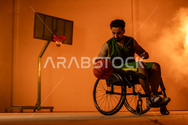 التدرب على إتقان لعب كرة السلة، أنشطة ترفيهية ورياضات شبابية، تحسين حياة ذوي الإعاقة الحركية، المحافظة على اللياقة البدنية والليونة، بورتريه لشاب عربي خليجي سعودي يرتدي الملابس الرياضية يجلس على كرسي متحرك يمسك الكرة بيده، اتباع نمط حياة صحي، خلفية برتقالية