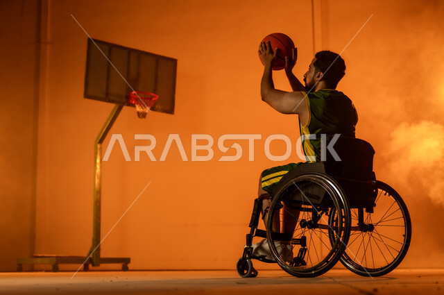 Recreational activities and youth sports, training to master playing basketball, improving the lives of people with motor disabilities, maintaining physical fitness and flexibility, portrait of a young Saudi Arabian Gulf man wearing sportswear sitting in a wheelchair holding a ball in his hand, following a healthy lifestyle, orange background