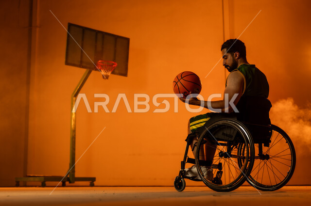 تحسين حياة ذوي الإعاقة الحركية، أنشطة ترفيهية ورياضات شبابية، التدرب على إتقان لعب كرة السلة، المحافظة على اللياقة البدنية والليونة، بورتريه لشاب عربي خليجي سعودي يرتدي الملابس الرياضية يجلس على كرسي متحرك يمسك الكرة بيده، اتباع نمط حياة صحي، خلفية برتقالية
