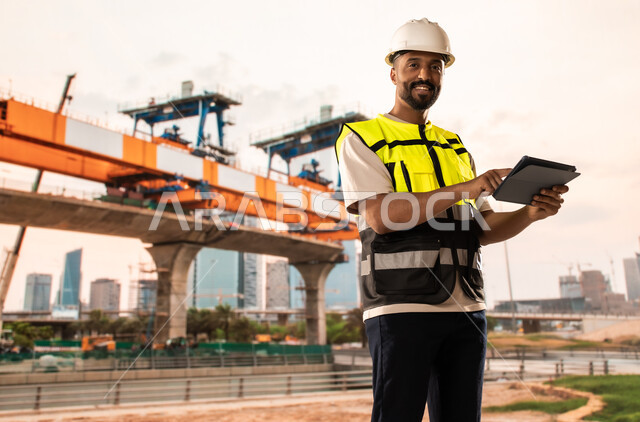 Integrating modern and advanced technologies into engineering work, working in the engineering sector, development and growth of the engineering field in the Kingdom of Saudi Arabia, a Saudi Arabian Gulf engineer wearing a protective jacket and helmet stands at the workplace and holds a tablet in his hands
