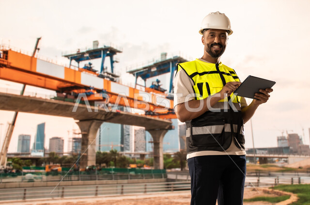 Integrating modern and advanced technologies into engineering work, working in the engineering sector, development and growth of the engineering field in the Kingdom of Saudi Arabia, a Saudi Arabian Gulf engineer wearing a protective jacket and helmet stands at the workplace and holds a tablet in his hands