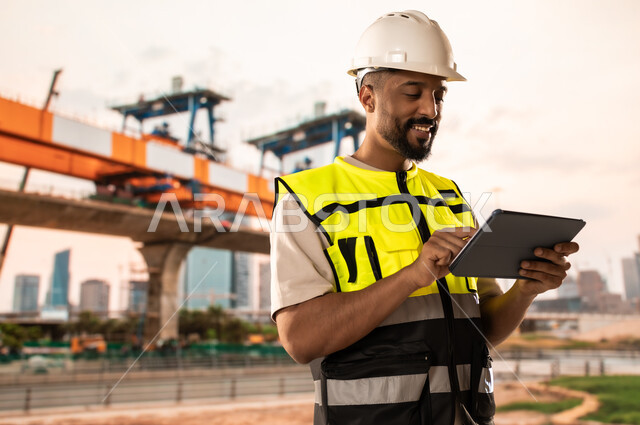 Integrating modern and advanced technologies into engineering work, working in the engineering sector, development and growth of the engineering field in the Kingdom of Saudi Arabia, a Saudi Arabian Gulf engineer wearing a protective jacket and helmet stands at the workplace and holds a tablet in his hands