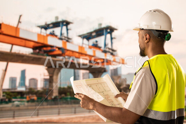 دراسة أساسيات المخطط الإنشائي, العمل القطاع الهندسي, مهندس عربي خليجي سعودي يرتدي سترة الحماية والخوذة يمسك مخططات الإعمار بيديه, مراجعة وعرض مخطوطات المشاريع بشكل كامل, إدارة ومتابعة المشروع، مفهوم الهندسة والانشاءات المعمارية