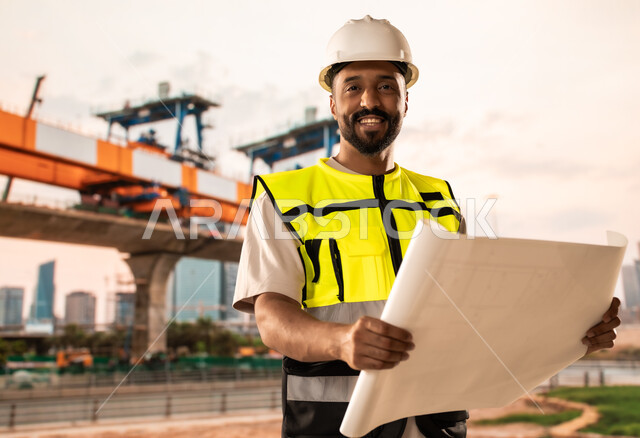 دراسة أساسيات المخطط الإنشائي, العمل القطاع الهندسي, مهندس عربي خليجي سعودي يرتدي سترة الحماية والخوذة يمسك مخططات الإعمار بيديه, مراجعة وعرض مخطوطات المشاريع بشكل كامل, إدارة ومتابعة المشروع، مفهوم الهندسة والانشاءات المعمارية
