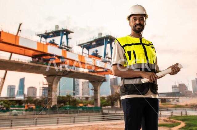 دراسة أساسيات المخطط الإنشائي, العمل القطاع الهندسي, مهندس عربي خليجي سعودي يرتدي سترة الحماية والخوذة يمسك مخططات الإعمار بيديه, مراجعة وعرض مخطوطات المشاريع بشكل كامل, إدارة ومتابعة المشروع، مفهوم الهندسة والانشاءات المعمارية